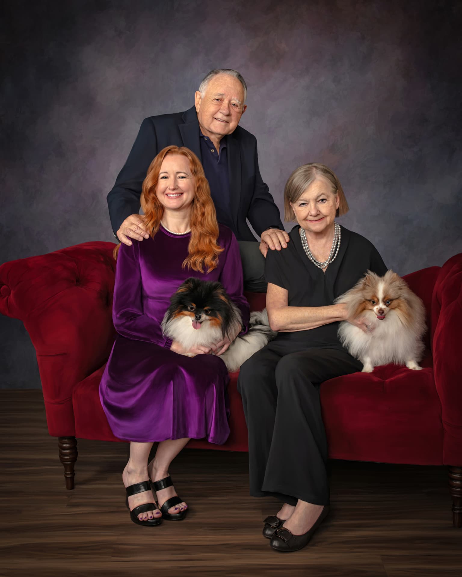 Studio portrait of Suzie, Frank, and Martie Miller on a red velvet couch with their Pomeranians Sky and Indy